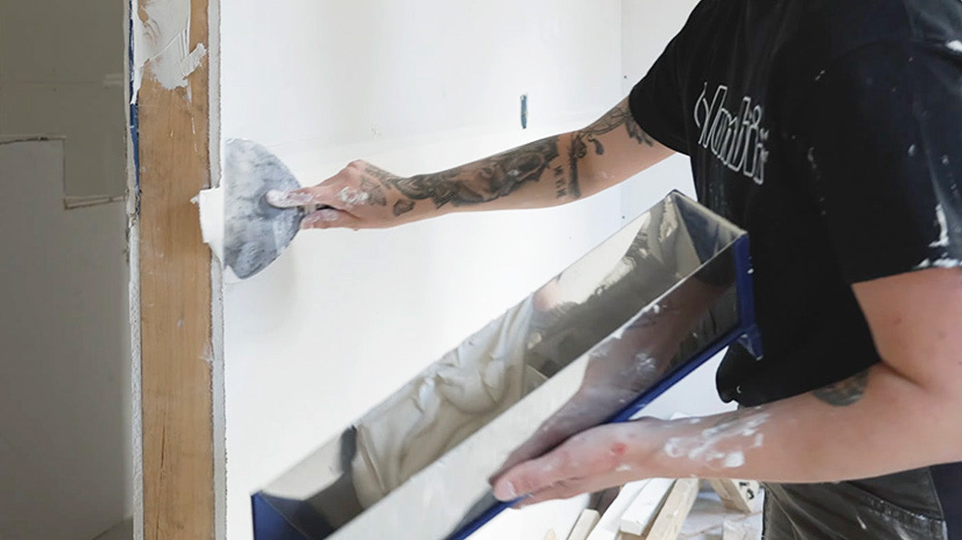 Drywaller applying joint compound over drywall tape with a joint knife while holding a mud pan